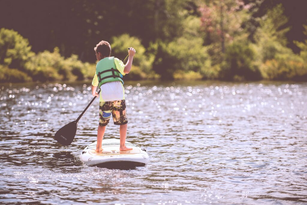 Ein Kind in Schwimmweste paddelt auf einem Board stehend über einen See.