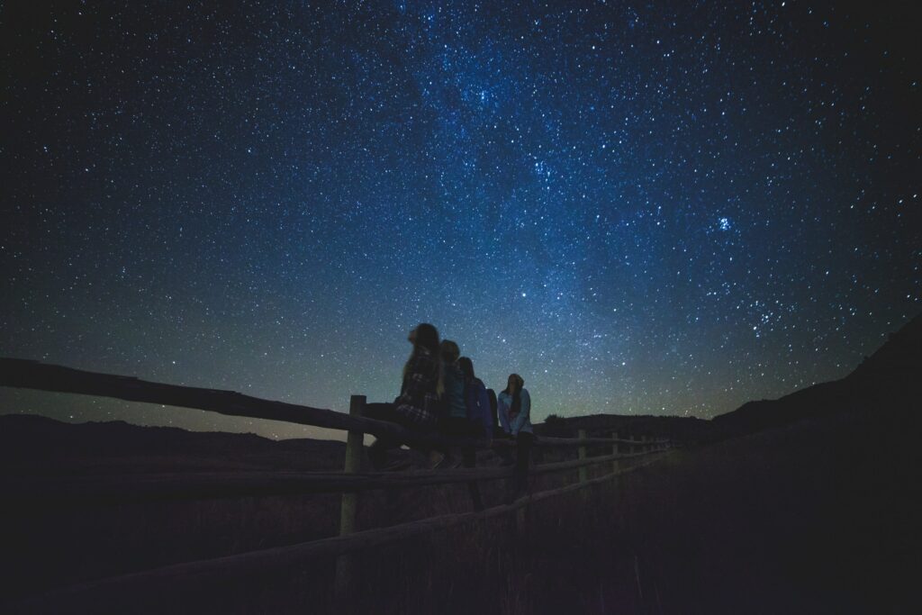 Menschen sitzen auf einem Holzzaun und schauen nach oben Richtung Sternenhimmel.