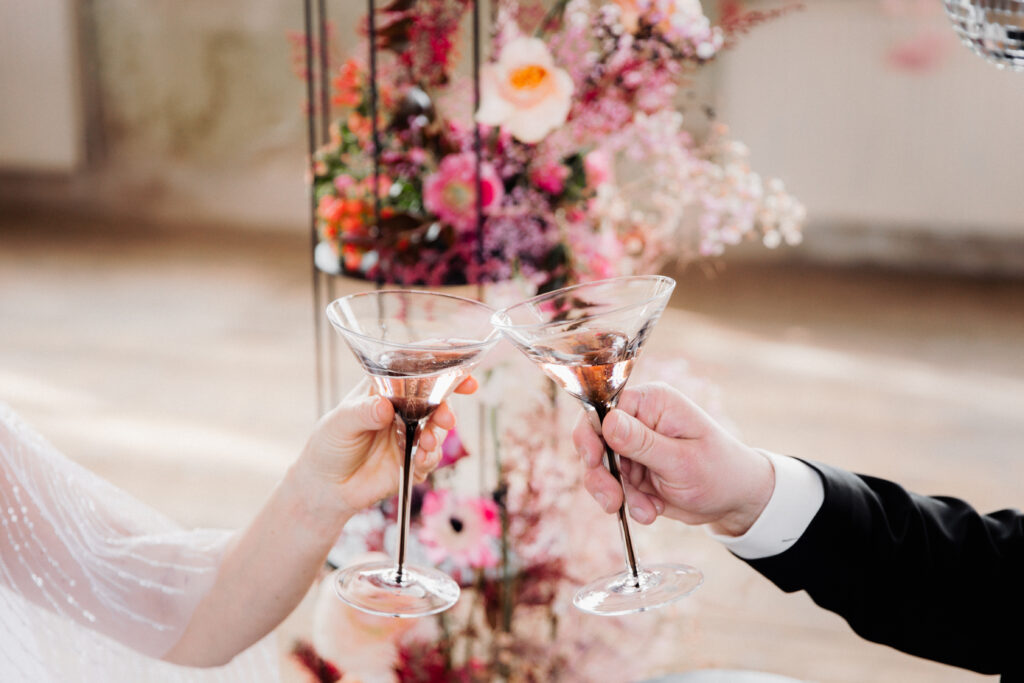 Ein Brautpaar stößt mit Sektschalen vor einem farbenfrohen Blumenarrangement an.