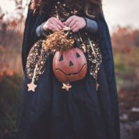 Kind steht verkleidet auf einem Feld und hält einen Halloween-Kürbis in der Hand