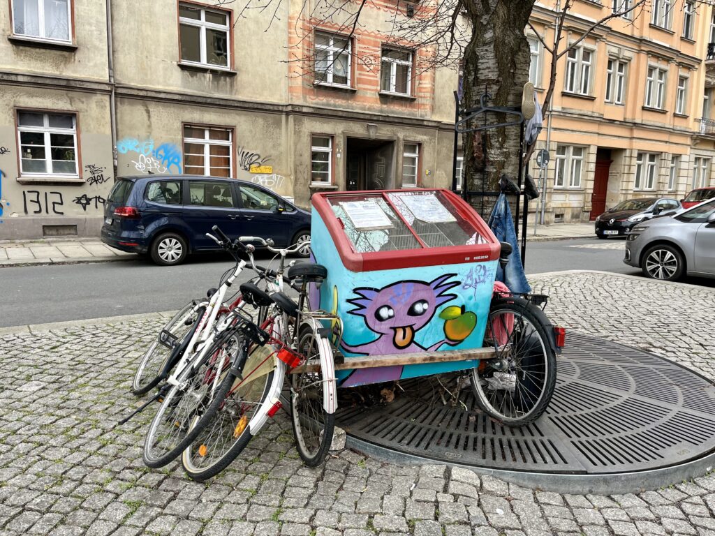 Eine alte Eistruhe mit rotem Deckel und kreativen Figur drauf gemalt – ein Fahrrad-Fairteiler in Dresden Pieschen