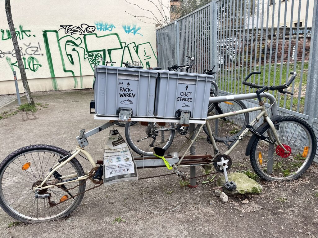 Fahrrad-Fairteiler: ein selbst zusammengebautes Fahrrad mit zwei grauen Kisten, für jeweils Gebäck und Obst, zur Abgabe und Weitergabe an andere