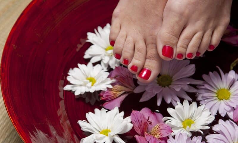 Füße mit rotem Nagellack über einer Schüssel mit Wasser und bunten Blüten