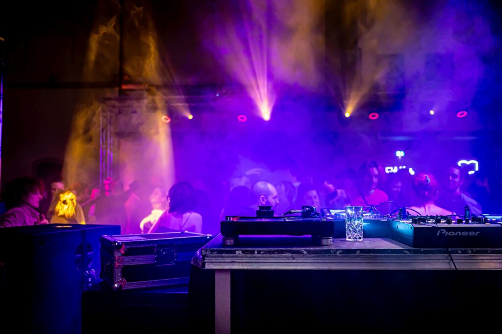 Turntables stehen auf einem Tisch, im Hintergrund tanzen Menschen in einem lila-blauen Licht, Nebel ist zu sehen